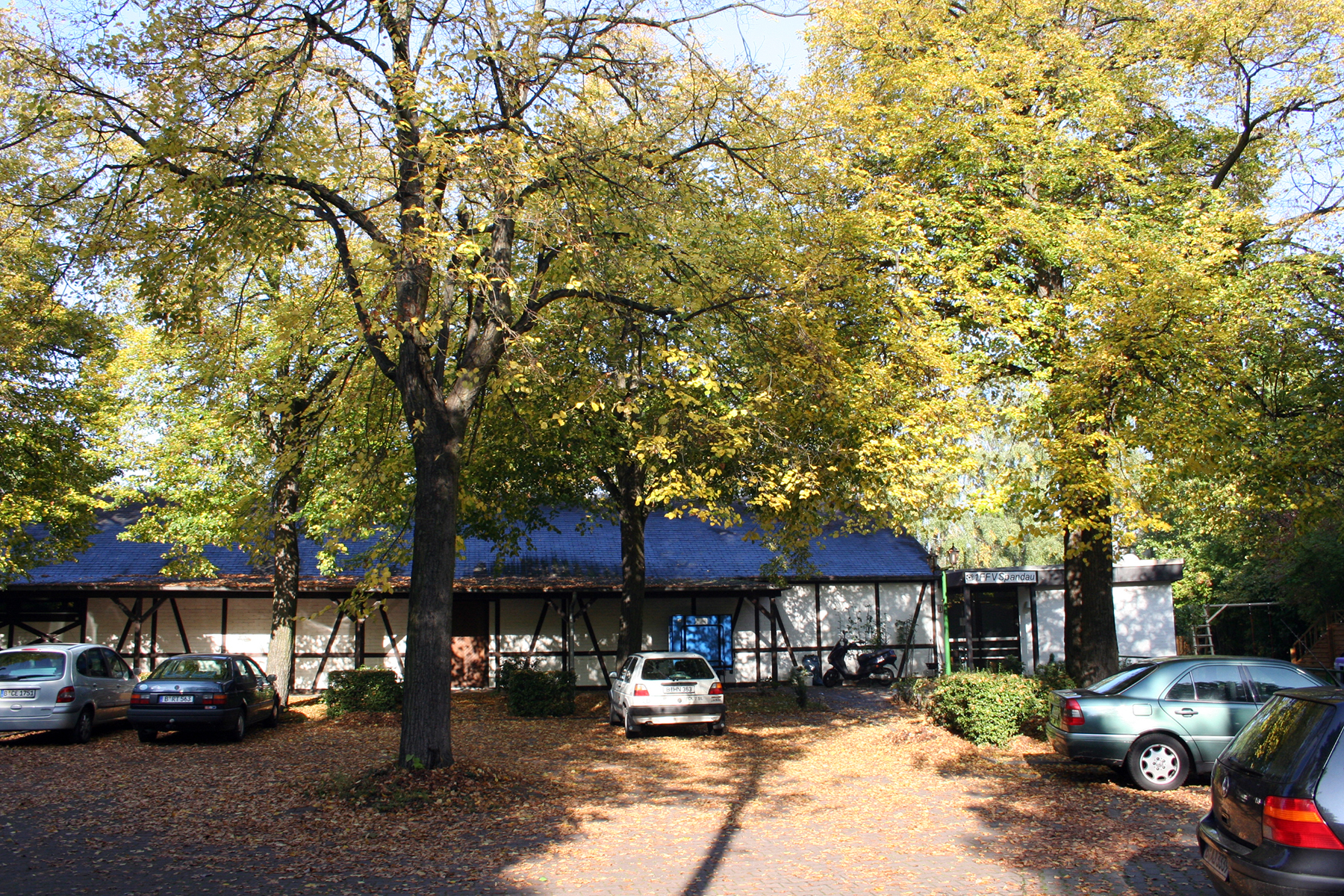 Einfahrt auf den Parkplatz. Mittig ein Baum, Blick auf die Seite der Schiesshalle, vorwÃ¤rts Richtung Hallenseite geparkte PKW. Rechte Seite der Halle, sowie in der Mitte, ein Eingang in das GebÃ¤ude und auf den SchieÃŸstand. Links oben ein schmales Fenster, dicht unter dem Dach.